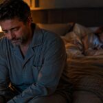 Hombre triste sentado en el borde de la cama mientras su mujer, acostada, mira el móvil con indiferencia: mi mujer no me desea